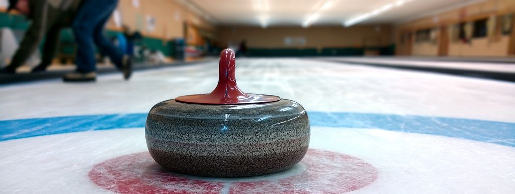 Curling stone with a red handle at rest on the button of an indoor sheet, players blurred behind